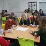 Students and a teacher sit around a table with drinks and paper at a workshop.