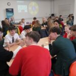 Students sit in a circle around a table, taking part in a group activity. Other groups and Citizens UK branding are visible across the room.