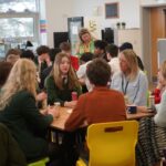 A group of students sits around a table (drinks are on the table). Additional discussion groups and facilitators are visible in the background near tall windows.