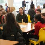 A group of students sits around a table (paper and drinks are on the table). Additional discussion groups and facilitators are visible in the background near tall windows.