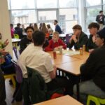 A facilitator stands beside a table of students who are seated in discussion. Drinks and worksheets are spread across the tables. More groups are visible in the background near large windows.