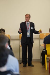 A man dressed in a suit is doing a speech to students.