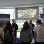 A group of people stand at a table with banners for "Summerfield Commercial" and "Summerfield Homes" in a brightly lit room with large windows. The scene appears to be a networking or informational event.