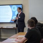 A man dressed in a suit is standing at the front of a classroom next to a screen, he is presenting.