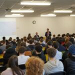 A group of people seated in a room facing a presentation screen that reads "Real Estate Exhibition." Three individuals appear to be presenting at the front, while the audience listens attentively. The setting is a classroom or lecture hall with a clock on the wall.