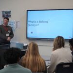 A person stands at the front of a classroom next to a large screen displaying the text "What is a Building Surveyor?" Several people are seated, facing the screen. Classroom equipment is visible around the room.