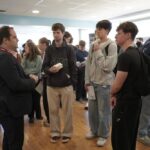 A group of people are gathered indoors in a networking setting, engaging in conversation. Some participants are holding papers or items. The room has large windows and wooden floors.