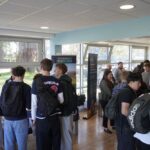 A group of people are gathered in a room with large windows, interacting around informational booths. Signs include "Summerfield Common Room" and "Summerfield House." Many individuals are wearing backpacks, and there are promotional materials on tables.