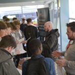 A group of people are gathered indoors around an information stand. Two individuals behind the stand are engaging with visitors.