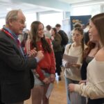 A group of people are engaged in conversation at an indoor event. An individual wearing a suit and a red lanyard appears to be talking to others who are casually dressed. The setting includes informational displays and windows in the background.
