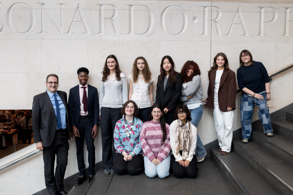 Articulation prize grand finalists stand with the adjudicators on steps at the national gallery. Behind them is part of some text that is carved into the wall.