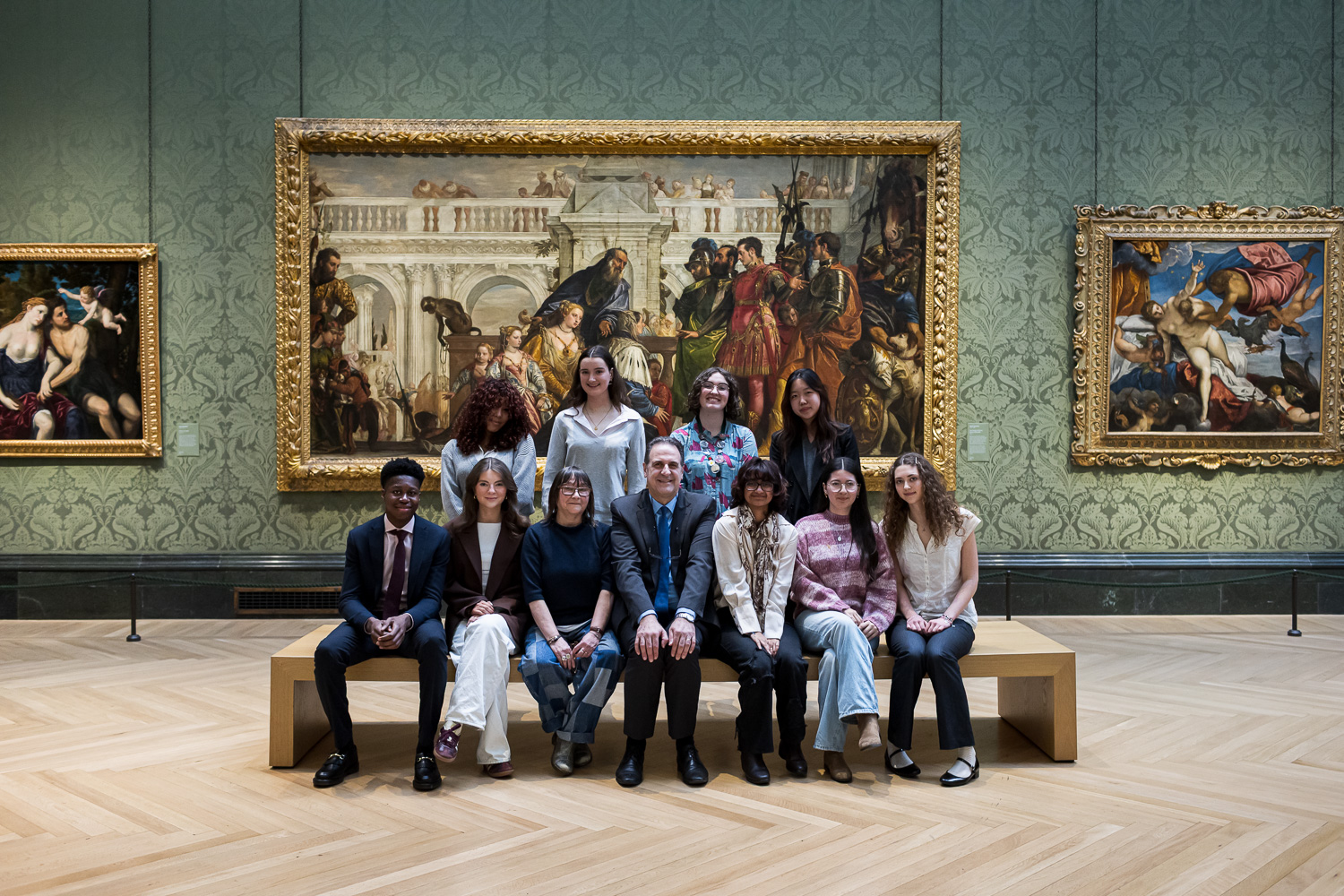 Articulation prize grand finalists sit with the adjudicators on a bench in the centre of a gallery in the national gallery. Behind them is a large painting in the middle of two smaller paintings.