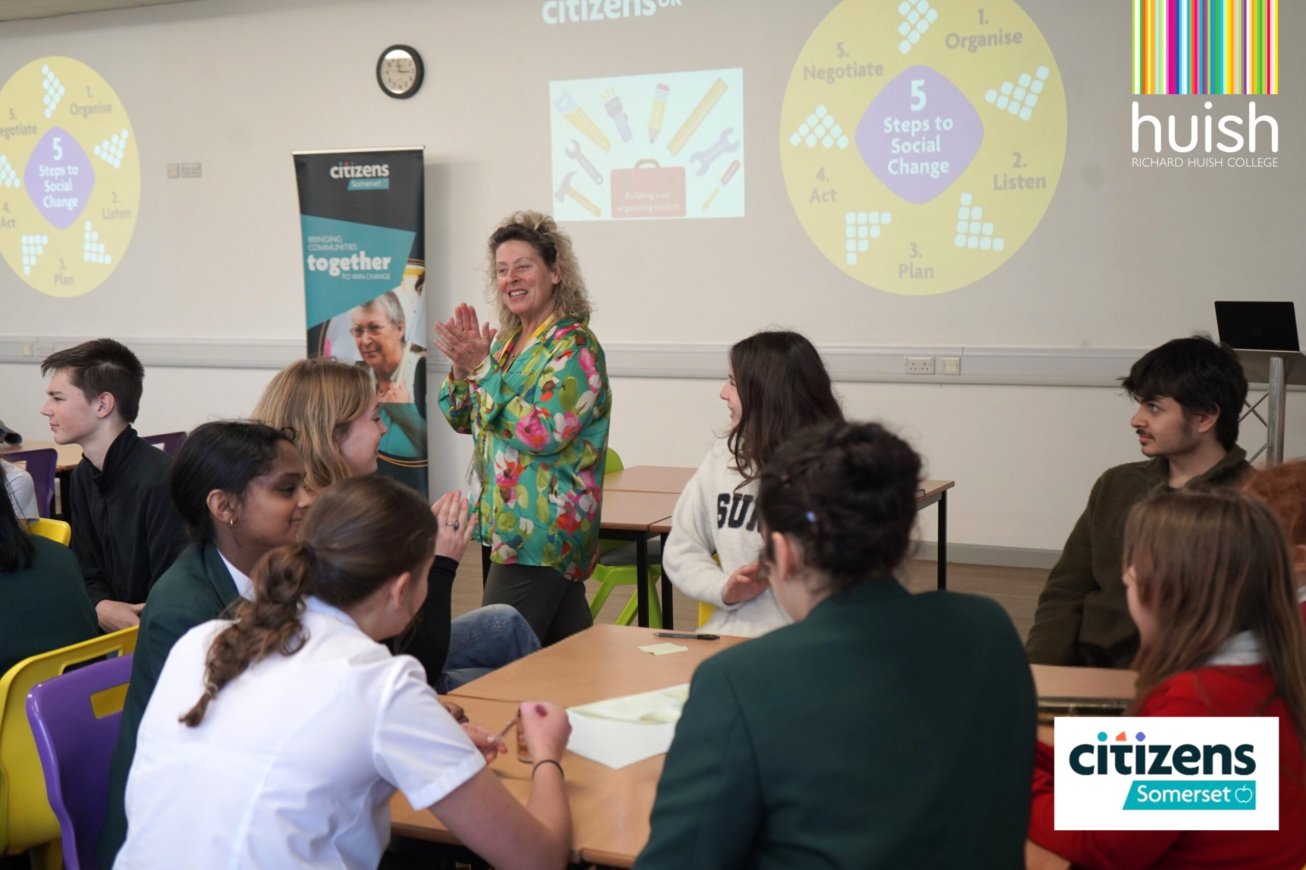 A facilitator speaks to a table of students. A projector displays Citizens UK content on the wall. Logos for Huish College and Citizens Somerset appear in the image.