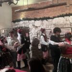 Participants dressed in traditional folk costumes are dancing in a cosy room with stone walls and rustic decor. A painting and several framed pictures are mounted on the walls, and a chandelier hangs from the ceiling.