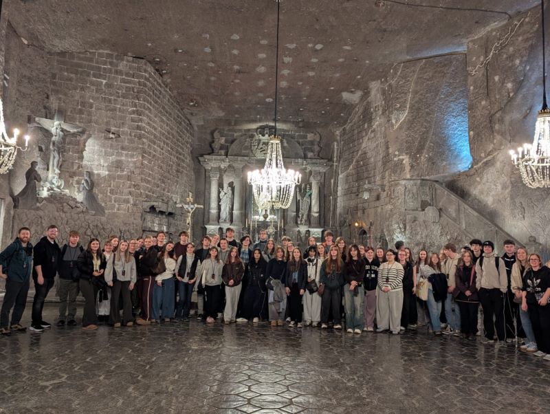 A large group of people stand together in the Wieliczka Salt Mine, surrounded by salt rock walls and ornate chandeliers. The environment is dimly lit, highlighting the intricate carvings in the salt.