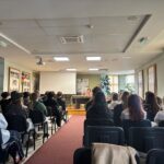 A group of people seated in a conference room facing a Holocaust survivor who is speaking to the room. The room has posters on the walls and yellow blinds. Masks can be seen on some members of the audience.