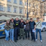 A group of people stand closely together in front of a police car on a city street in Poland. An officer is amongst them, and the background features buildings with signs reading "Metropolitan" and "Exchange.