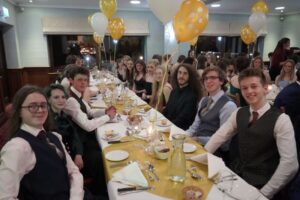 A group of students sat at a table. The table is set in front of them and they are dressed formally. Yellow and white balloons are placed in various places on the table.