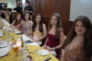 A group of students sat at a table. The table is set in front of them and they are dressed formally.