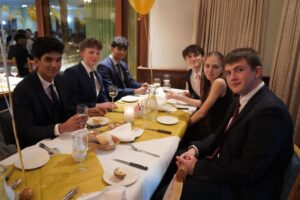 A group of students sat at a table. The table is set in front of them and they are dressed formally.