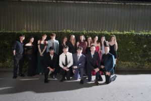 A group of students in front of a hedge. Some are stood and those on the front row are kneeling on one leg. They are dressed formally in ball gowns and suits.