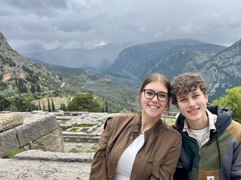 Two people sitting together and smiling with a dramatic mountain landscape behind them at Delphi. The sky is cloudy and the ancient stone site is partly visible in the foreground.