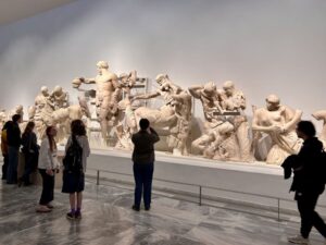 Museum visitors standing in front of a long display of large ancient Greek marble sculptures depicting a dynamic battle scene. The statues are arranged against a plain white wall in a well‑lit gallery.