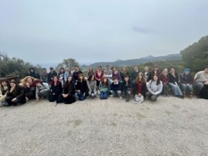 A large group of people posing together outdoors on a gravel area with hills and trees in the background. The weather is overcast, and the group is seated and standing in a wide formation.