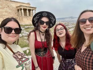 Four people smiling and posing for a selfie at the Acropolis in Athens, with the Erechtheion temple visible in the background. They are standing among ancient stone ruins under an overcast sky