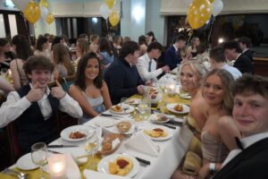 A group of students sat at a table. The table is set in front of them and they are dressed formally.