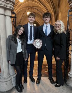 Four Huish Law School students stand in an arched corridor inside the Royal Courts of Justice, smiling and holding the National Bar Mock Trial shield after winning the competition.