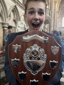 A Huish student in a suit holding the National Bar Mock Trial trophy close to the camera, smiling with visible excitement inside the Royal Courts of Justice.