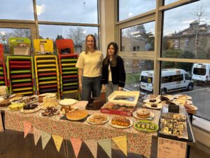Two students stand behind a table full of cakes. There is bunting on the front of the table that reads 'cake sale'. 