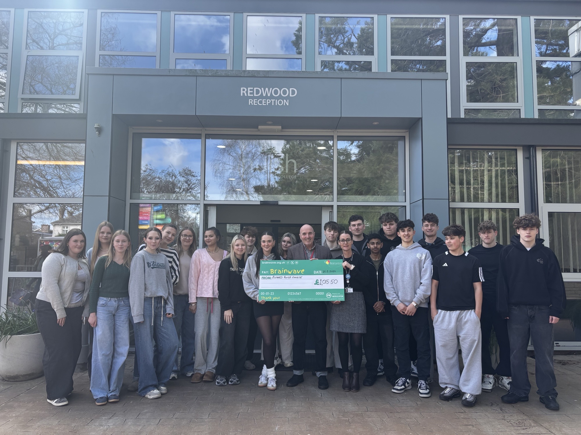 A group of people stand in front of the college reception. They are holding a large cheque for Brainwave.