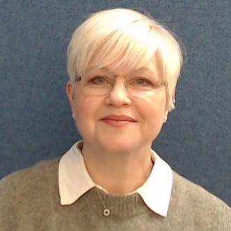 A lady with short blonde hair and glasses smiles at the camera. She is against a blue backdrop and wears a white shirt, green/grey jumper and silver necklace.