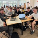 5 students sat around a table with laptops and paper on. They are smiling and one is doing a thumbs up.