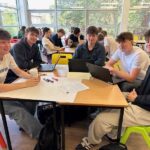 4 students sat at a table smiling, with laptops and posters they have written on in front of them.