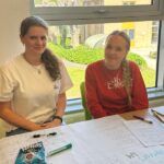 2 students sat at a table smiling, with posters they have written on in front of them.