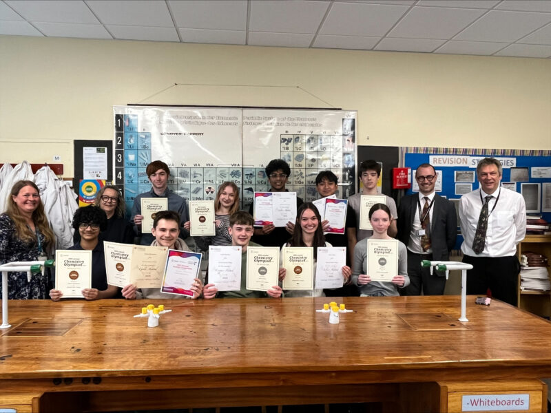 Students and staff standing and sitting in a lab. The students are holding their olympiad certificates.