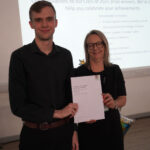 A prize-winning student stands with Emma Fielding, they are holding the student's certificate and smiling.