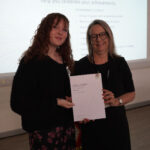 A prize-winning student stands with Emma Fielding, they are holding the student's certificate and smiling.