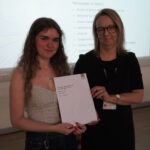 A prize-winning student stands with Emma Fielding, they are holding the student's certificate and smiling.