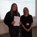 A prize-winning student stands with Emma Fielding, they are holding the student's certificate and smiling.