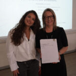A prize-winning student stands with Emma Fielding, they are holding the student's certificates and smiling.