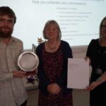 A prize-winning student stands with a sponsor and Emma Fielding, they are holding the student's certificate and trophy, and are smiling.