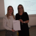 A prize-winning student stands with Emma Fielding, they are holding the student's certificate and smiling.