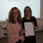 A prize-winning student stands with Emma Fielding, they are holding the student's certificate and smiling.