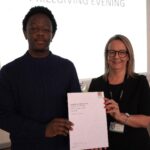 A prize-winning student stands with Emma Fielding, they are holding the student's certificate and smiling.