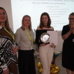 Sponsor representatives stand with a prize-winning student who is holding her certificate and trophy. Emma Fielding claps beside them.