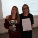 A prize-winning student stands with Emma Fielding, they are holding the student's certificate and trophy, and are smiling.